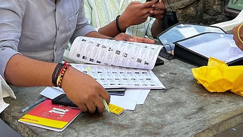 A group of people gathers around a table outside. One person is holding a booklet with rows of images and text, directory containing names of the voters in the November 2025 bypolls. Nearby are papers, a phone, and a yellow bag on the table.