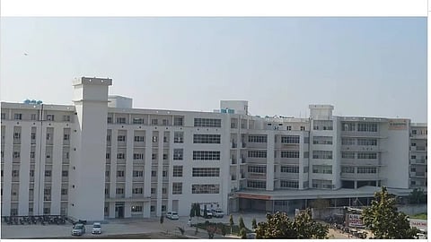 A large, modern multi-story building with numerous windows under a clear sky. Several parked cars are visible in front, suggesting a commercial or institutional facility. Trees and greenery are in the foreground.