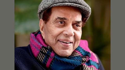 Dharmendra smiling warmly, wearing a tweed cap and a colorful scarf with patterns. The background is softly blurred, suggesting an outdoor setting.