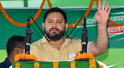 tejashwi yadav in a beige shirt speaks passionately at an event, raising one hand. The setting is decorated with orange marigold garlands, conveying a vibrant atmosphere.