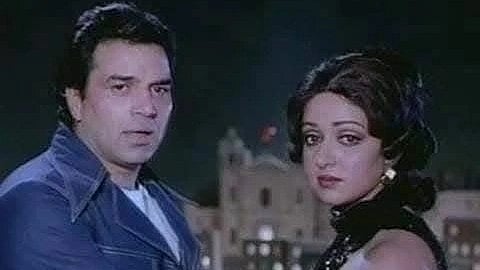 A man and woman stand closely under a night sky, with a building in the background. The mood is dramatic, and both appear contemplative.