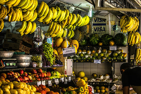 Fruit market stall with vibrant arrangement of hanging bananas, watermelons, and grapes. A person is seen attending to the colorful display.