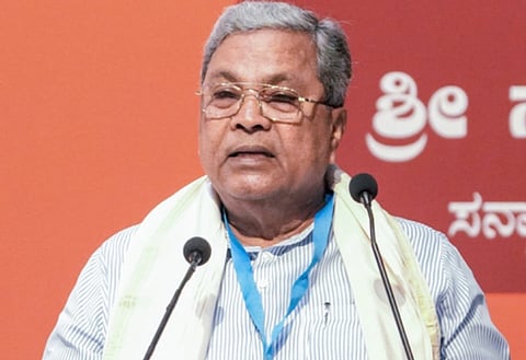 Minister for Labour Santosh S. Lad  wearing glasses speaks at a podium, with a microphone in front of him. The background is orange with text in a regional script.