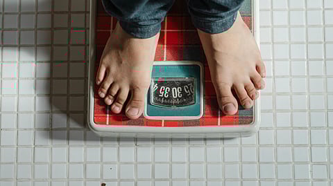Bare feet on a red plaid bathroom scale, displaying a weight in kilograms. The tiled floor suggests a home or bathroom setting.