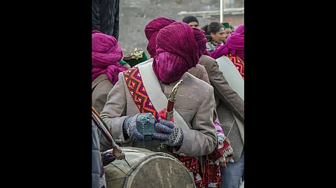 A person in traditional attire with a magenta fabric covering their face, he even has a knife in his hand. They wear gloves and a decorative sash, surrounded by others in similar outfits for the Raulane mela.