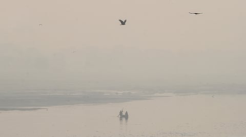 A thick morning haze blankets the river as a boat floats on a cold morning amid rising air pollution. There are even two birds flying in the sky.