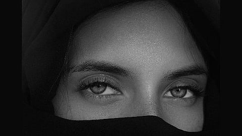 A close-up of a person with olive green eyes, partially covered in a black headscarf. The expression is calm and mysterious, with soft lighting.
