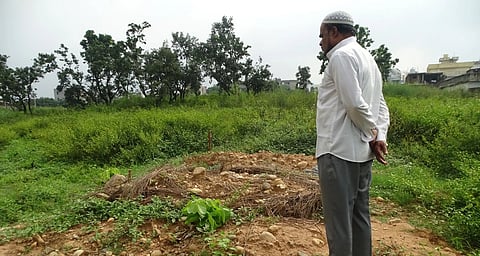 A man stands in front of a grave in a field. he is wearing a white te hsirt, grey pants, and a skull cap.