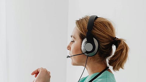 A healthcare professional in green scrubs sits at a desk, using a headset and laptop for a video call. A calm, focused atmosphere prevails.