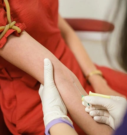 A healthcare professional wearing gloves draws blood from a patient's arm using a needle. The patient is in a red shirt, sitting calmly in a clinic setting.