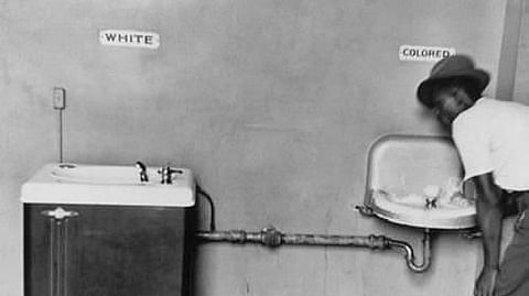 Black and white photo of segregated water fountains labeled "White" and "Colored." A man drinks from the "Colored" fountain, conveying racial segregation.