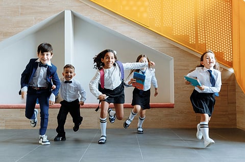 Students in uniform running together inside the school; girls wearing black skirts with white shirts and ties, and boys in black trousers with white shirts and ties. They are carrying school bags and notebooks.