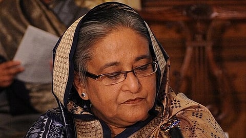 Sheikh Hasina wearing glasses and a patterned saree is sitting, giving a thoughtful look. The background is blurred, suggesting an indoor setting.