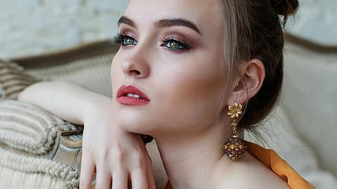 Young woman gazes thoughtfully, wearing floral gold earrings and an orange top. Her makeup is elegant, with smoky eyeshadow and pink lips, exuding sophistication.
