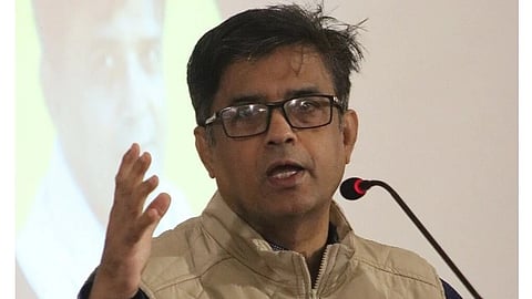 Bangladesh Press Secretary Shafiqul Alam speaking at a microphone, wearing glasses and a beige jacket, with a blurred portrait of himself projected in the background, conveying focus and engagement.