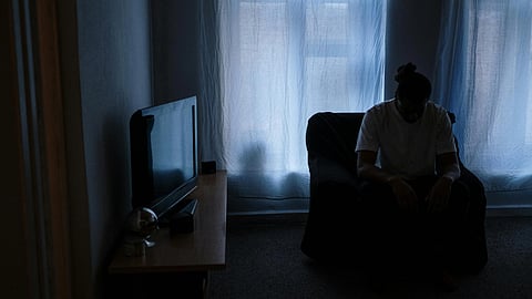 A person sits on a chair in a dimly lit room, silhouetted against sheer curtains with light filtering through. The scene conveys solitude and introspection.