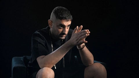 Haftbefehl sits on a dark chair against a black background, looking intently ahead with hands clasped. He wears a black t-shirt, chain, and bracelet, conveying a focused and serious tone.