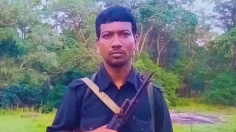 Madvi Hidma standing in a black uniform and a rifle in front of a forest.