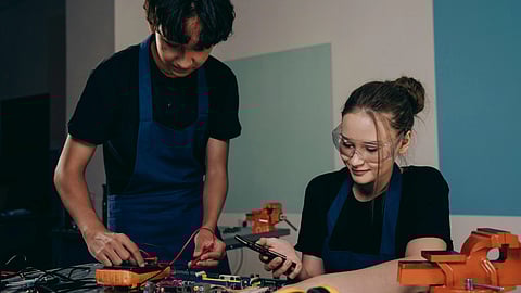 A girl and a boy in blue aprons work on an electronics project at a table. The young man focuses on wiring, while the young woman holds a tool, smiling slightly.