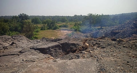 A pit on a stony mound with smoke rising from within, with field and trees in the background.