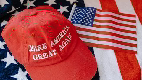 Red "Make America Great Again" hat and small American flag on an American flag backdrop. Patriotic theme, evoking national pride.