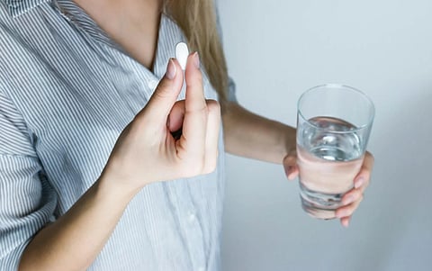 A person in a striped shirt holds a large white pill in one hand and a glass of water in the other, suggesting they are about to take medication.