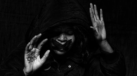 A grayscale photograph of a woman in a hoodie, her expression filled with fear and distress. Her mouth is tightly sealed with tape, and she raises her hands defensively. The word ‘STOP’ is written across her palms, amplifying her silent plea for help.