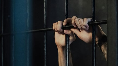 Hands gripping prison bars in a dimly lit cell, conveying a sense of confinement and desperation. The person wears a white shirt and has tattoos on their arms.