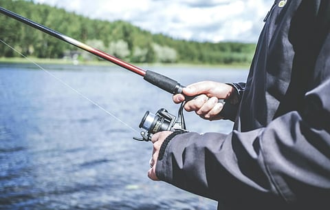Representational mage depicts a person standing by a tranquil lake, holding a fishing rod with a focused grip. The background shows a forest and cloudy sky, conveying a peaceful mood.