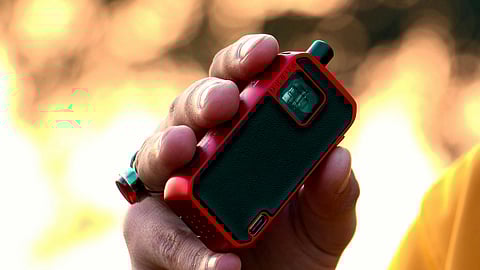 A hand in a yellow sleeve holds a small red and black vape, with blurred warm-toned bokeh lights in the background, creating a cozy ambiance.