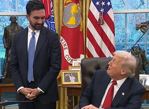 Image of Zohran Mmdani  in a suit standing and smiling beside Donald trump who is seated in the Oval Office. trump is looking up, set in an office with flags and a window. The tone is formal yet lighthearted.