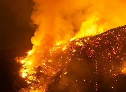 Representational image depicting a powerful wildfire engulfs a hillside at night, with intense orange flames and thick smoke billowing upwards, creating a dramatic and dangerous scene.