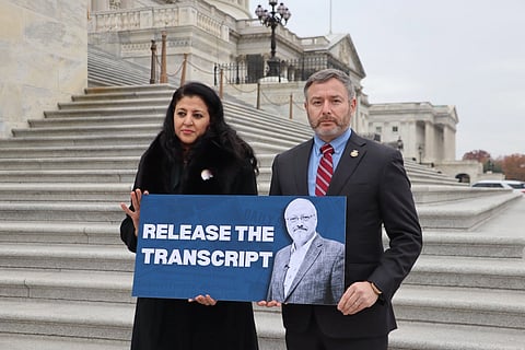 A congressman Eugene Vindman and a Khashoggi widow stand on the Capitol steps holding a sign reading "Release the Transcript" with an image of a bearded man, conveying urgency.