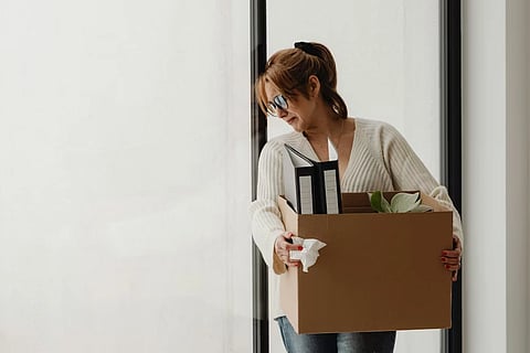 A woman standing next to a window, visibly sad. She is holding a box of office things likes files, implying she is fired.