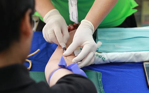A healthcare professional in gloves draws blood from a person's arm with a syringe. The patient wears a blue tourniquet. Medical supplies are visible on the table.