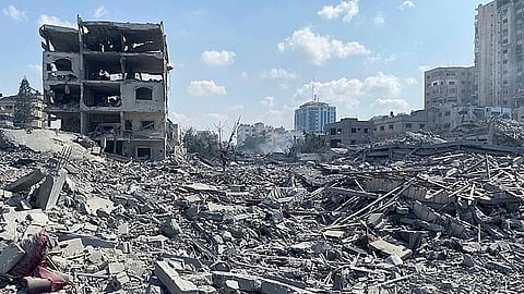 Rubble and debris fill the scene of a city in ruins under a clear blue sky, with a partially destroyed building on the left and damaged structures on the horizon.