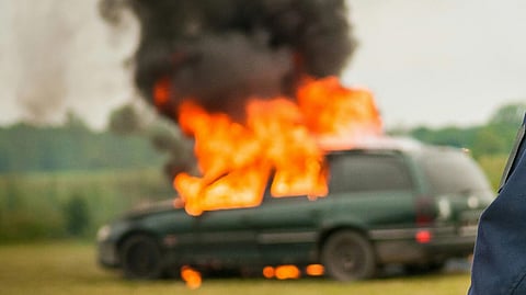  a car engulfed in bright orange flames and thick smoke in an open field.