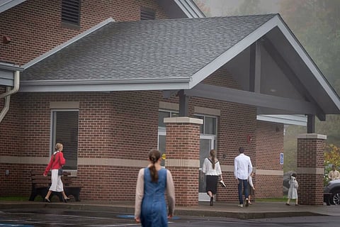 People walk toward a brick building with a gabled entrance on a foggy day, conveying a calm and reflective mood. Some wear casual, modest clothing.