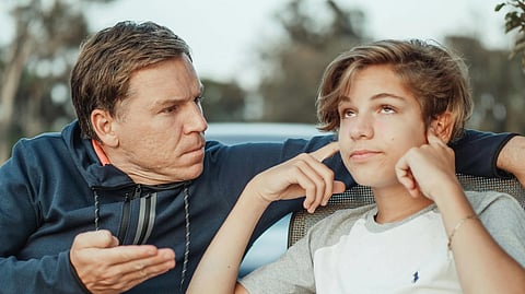 A man in a navy hoodie gestures while speaking to a boy in a white and gray shirt, who rolls his eyes and covers his ears, conveying frustration.