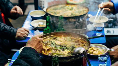 Steaming hot pot on a blue table, surrounded by people using chopsticks. Bottles, bowls of kimchi, and utensils evoke a lively, communal meal.