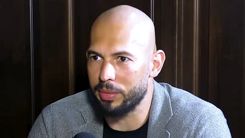 In the image Andrew Tate is shown talking to someone in a mic he is wearing grey coat and black t shirt