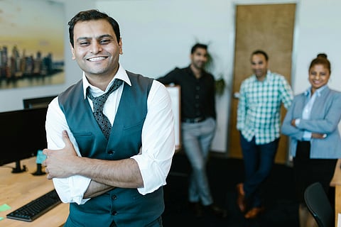 A man in a vest and tie smiles confidently in an office, with three colleagues blurred in the background. The atmosphere is friendly and professional.