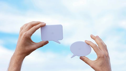 Two hands hold up white speech bubble cutouts against a bright blue sky with clouds, symbolizing communication and conversation.