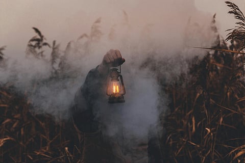 A person holding a glowing lantern in a misty field of tall grasses, creating a mysterious and moody atmosphere.