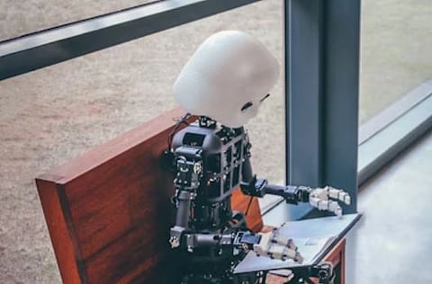 A robot sitting on a bench using a tablet with a glass wall behind it.