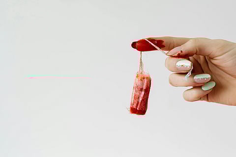 A hand with pastel nail polish holds a tampon covered in red paint against a plain background, symbolizing menstruation and conveying a bold, unapologetic tone.