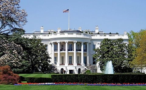 The White House with its iconic columns stands majestically under a clear sky, with an American flag atop. A fountain and colorful flowers adorn the foreground.