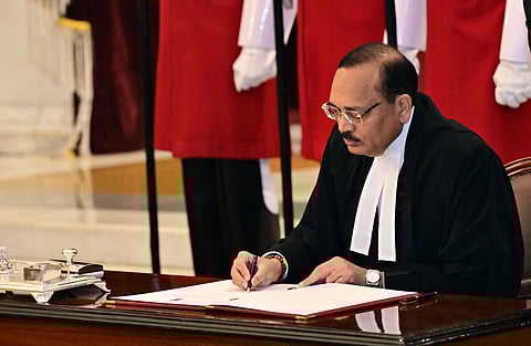 Image of justice Surya Kant in formal attire signs a document at a desk, surrounded by individuals in red robes. The scene is solemn, suggesting an official ceremony.