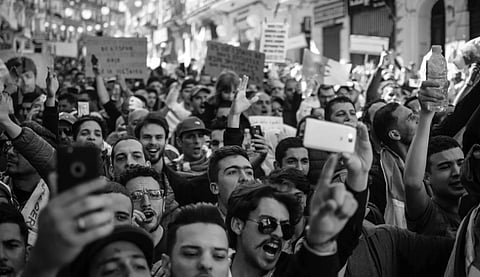 Black-and-white image of a large, energetic crowd of protesters holding signs and smartphones. The atmosphere is lively and passionate.