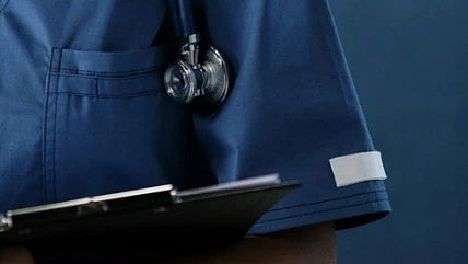 A healthcare worker in blue scrubs holds a clipboard with a stethoscope over their shoulder. The background is a matching blue, creating a professional tone.